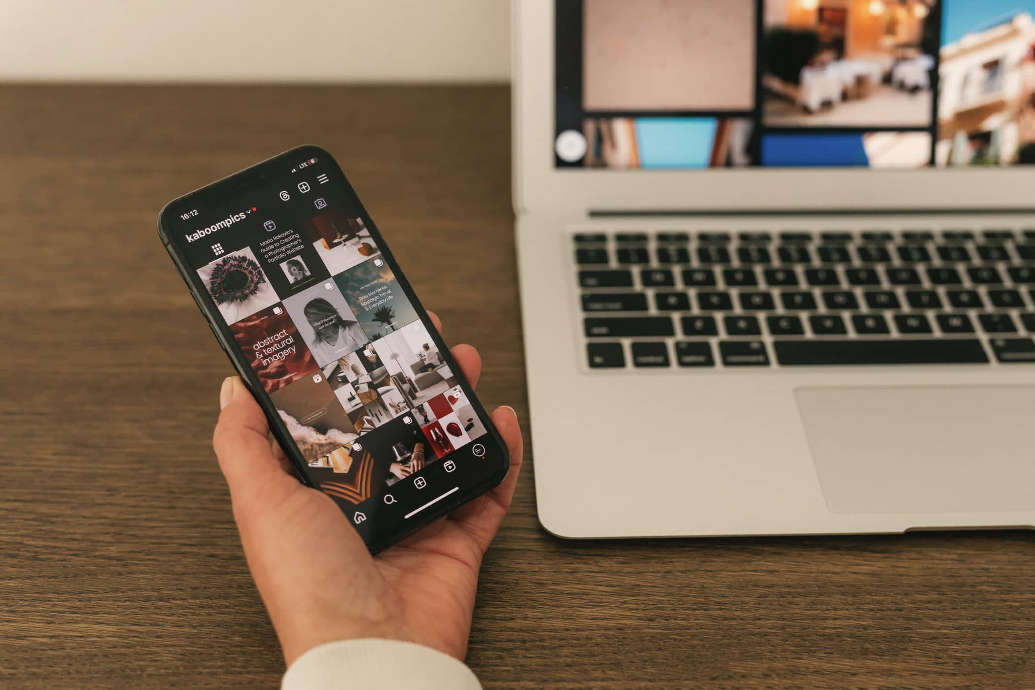 A hand holding a phone showing a social media feed beside an open laptop on a wooden desk