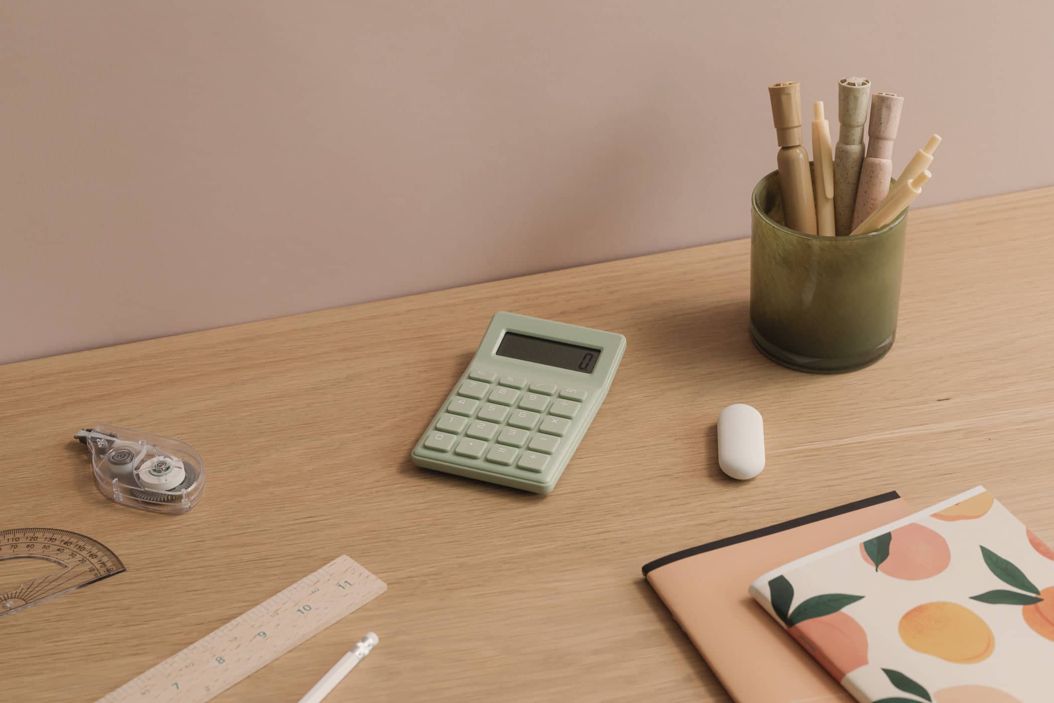 A sage-green calculator on a light oak desk beside notebooks, a ruler and a pot of pencils