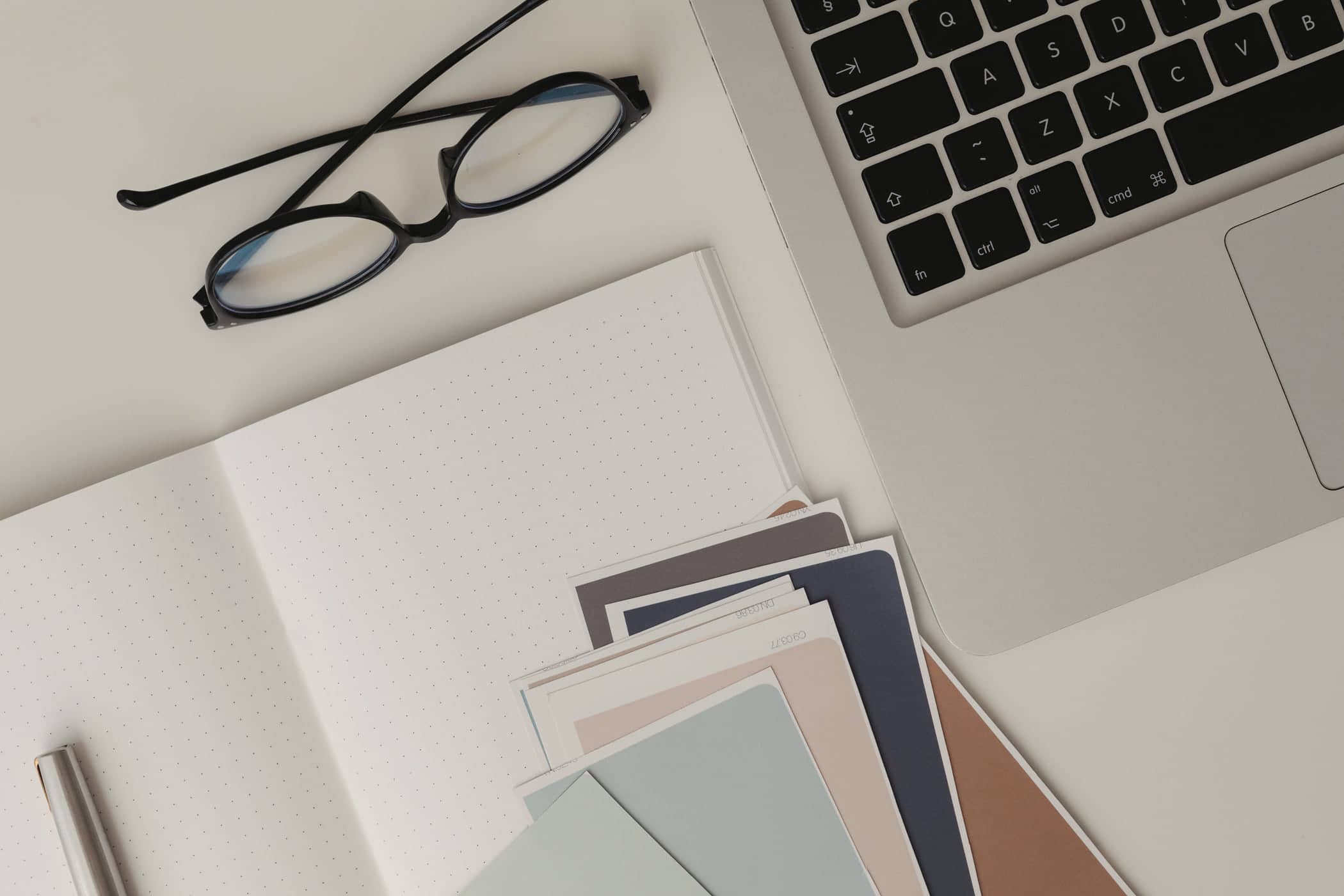 Paint colour swatches and a pair of glasses on a white desk beside a laptop
