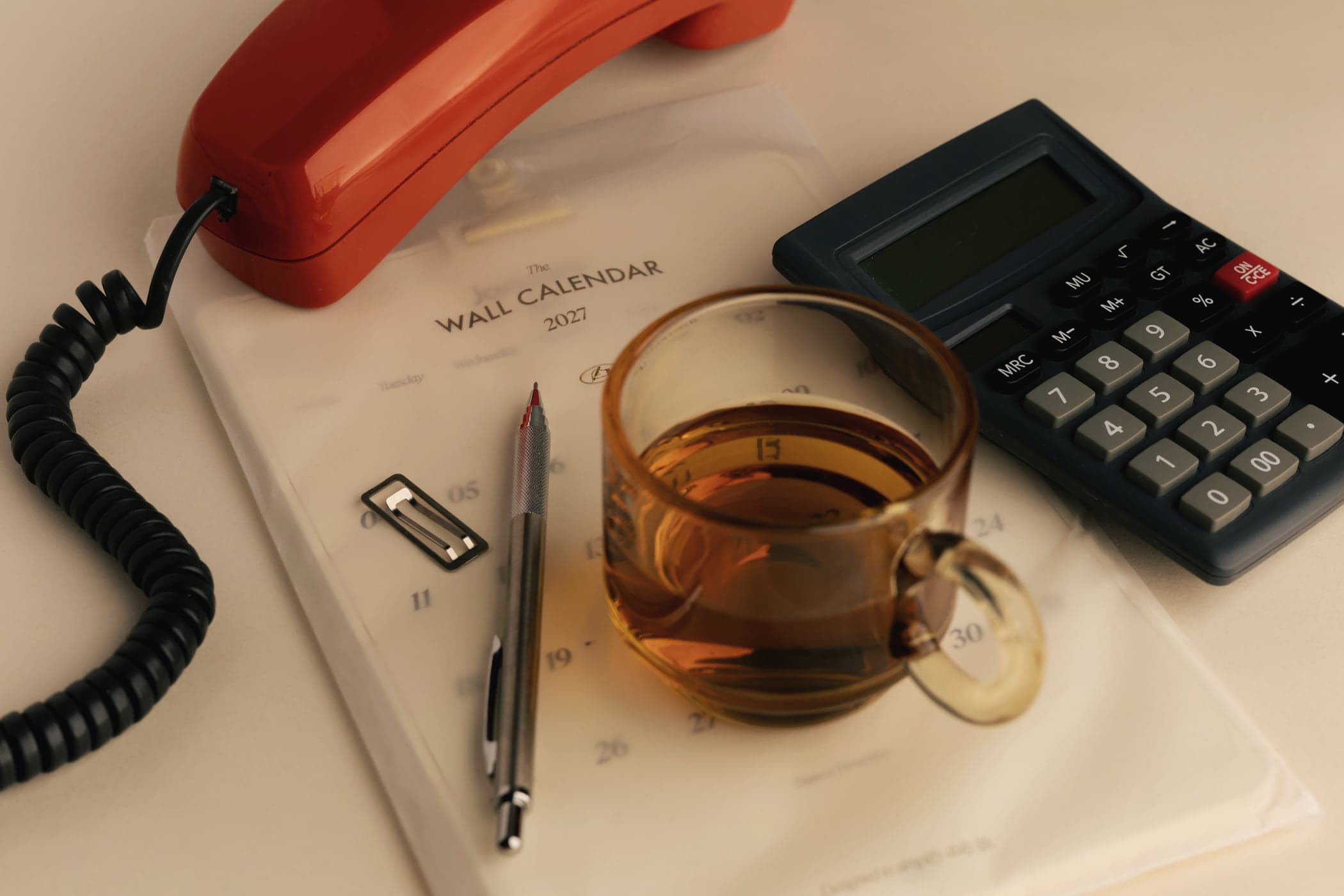 A retro telephone, a calculator, a wall calendar and a glass of tea on a desk
