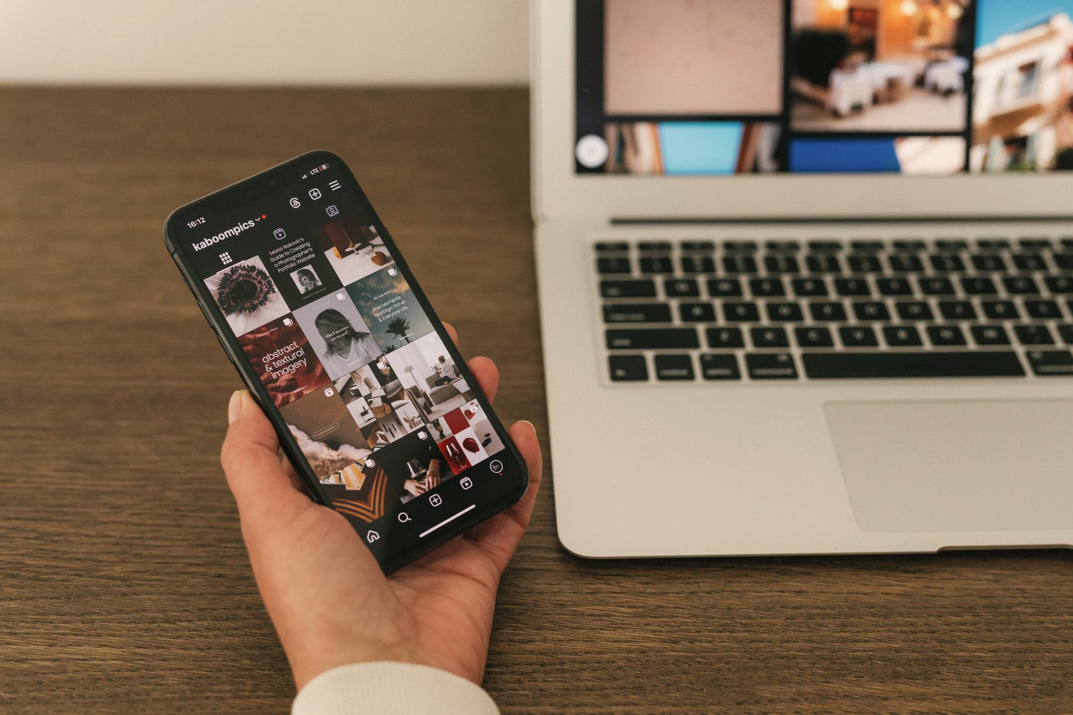 A hand holding a phone showing a social media feed beside an open laptop on a wooden desk