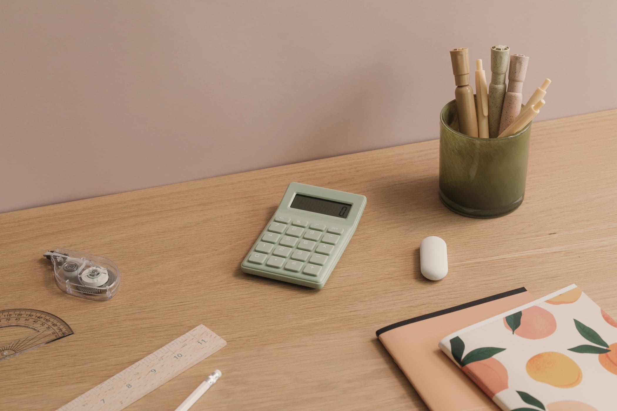 A sage-green calculator on a light oak desk beside notebooks, a ruler and a pot of pencils