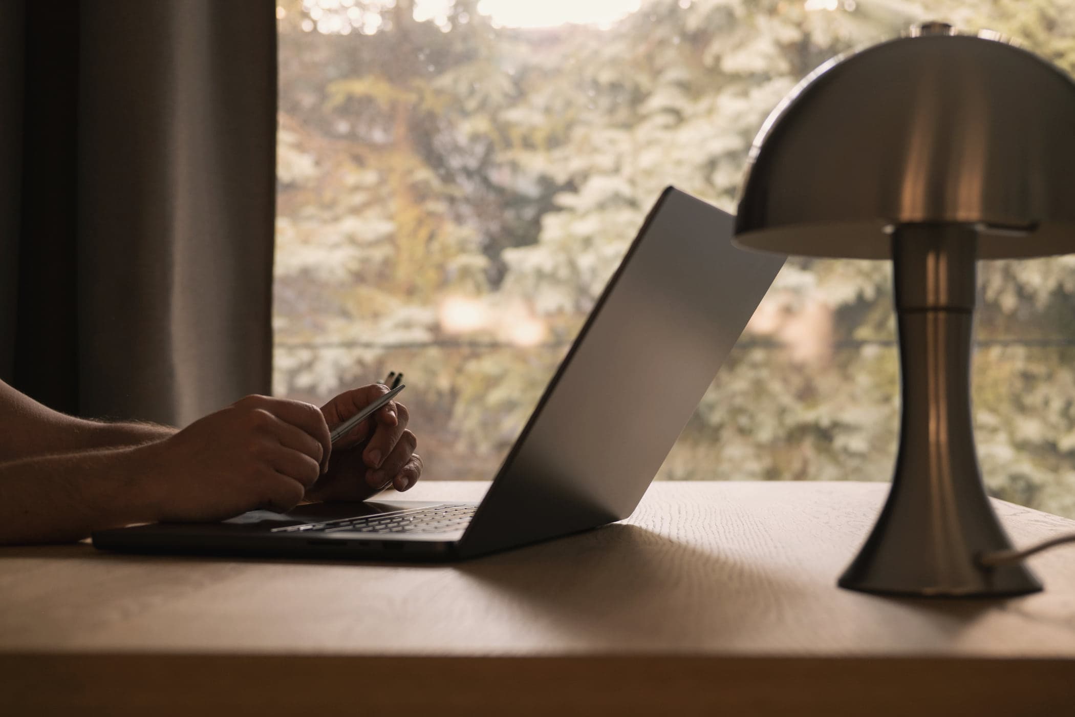 A laptop open on a warm oak desk by a window, a brass table lamp beside it