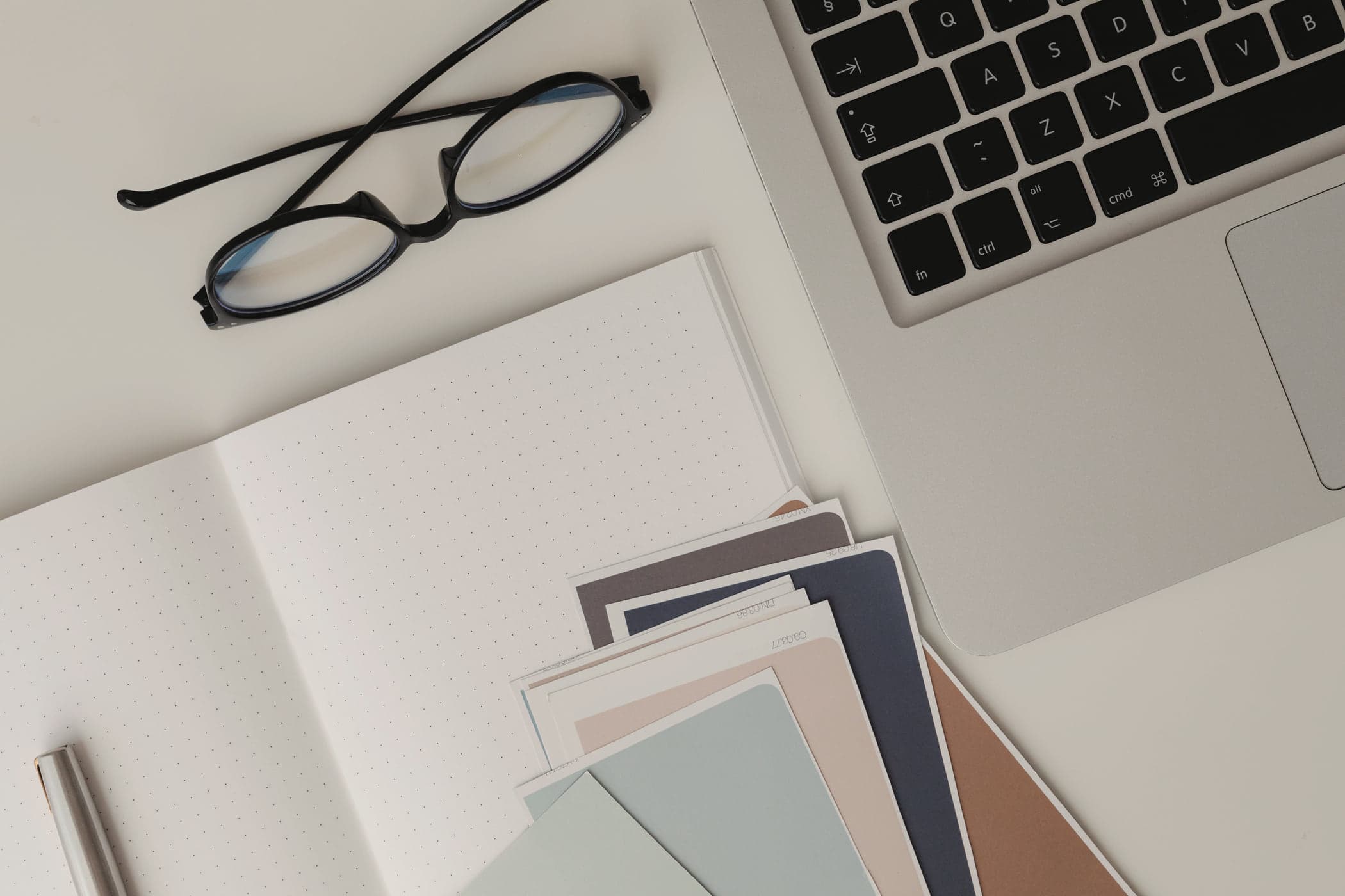 Paint colour swatches and a pair of glasses on a white desk beside a laptop